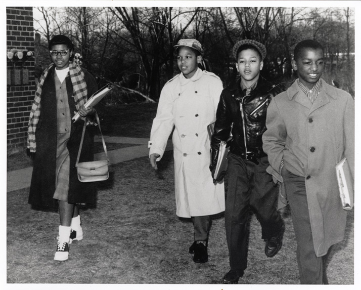 Four black students walk towards the camera, wearing winter coats. They are carrying books and lunch baskets.