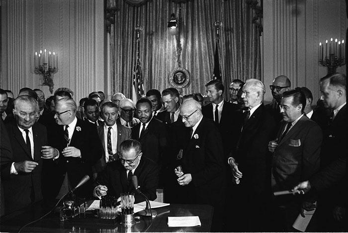 President Lyndon B. Johnson signs the Civil Rights Act of 1964 into law as various Civil Rights leaders look on, including the Reverend Martin Luther King, Jr. Photo courtesy of the LBJ Library, photo by Cecil Stoughton .