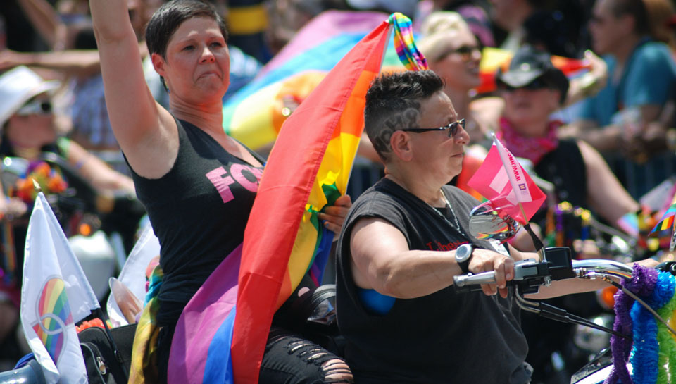 Photo of 2 women in black t-shirts carrying a rainbow flag and riding a motocycle in a Pride parade