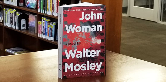 Photo of a book with a red and black cover with title "John Woman" in strong white font