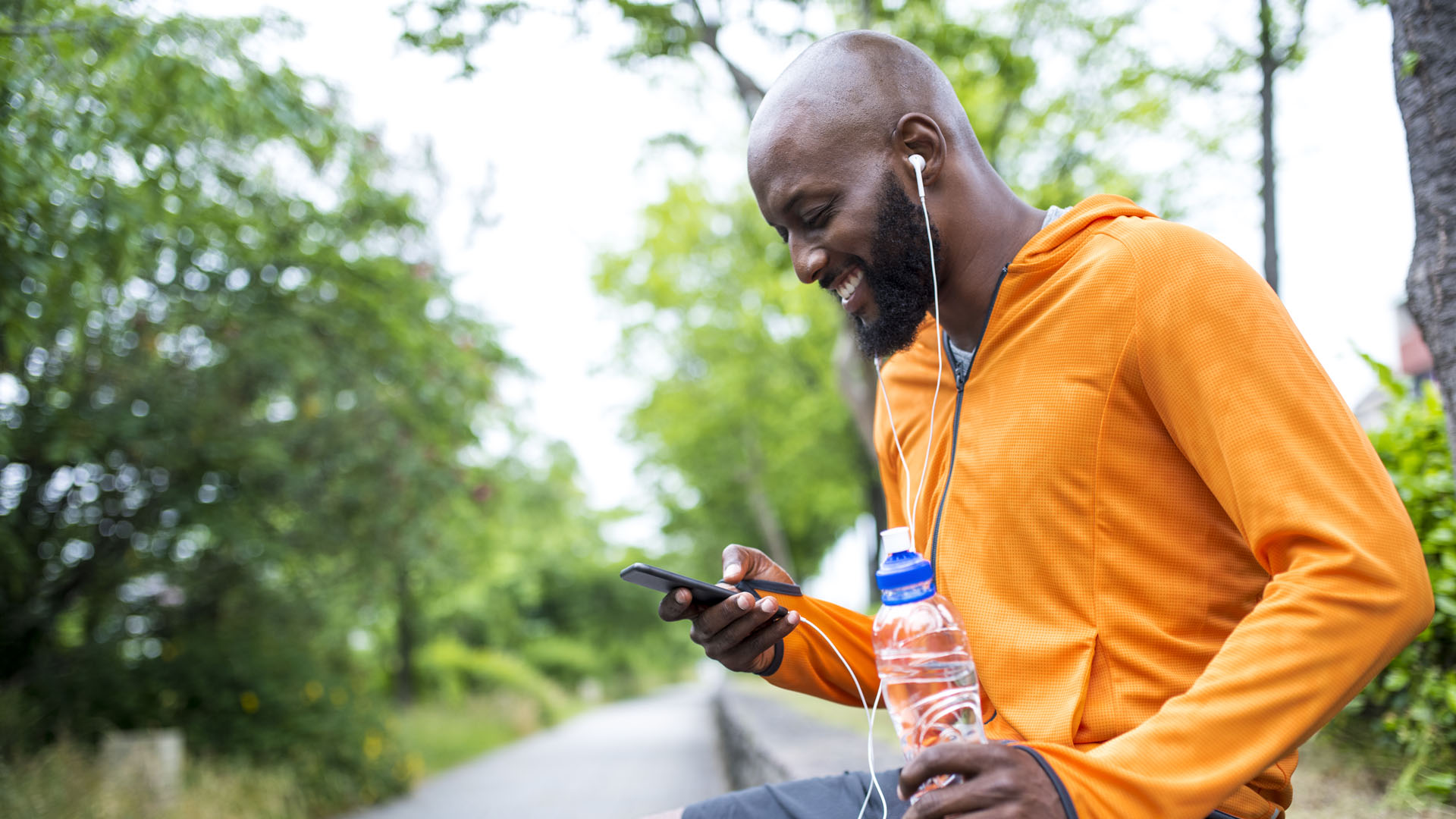 African american man sitting outdoors before his training. He is holding mobile phone in one hand and bottle of water in other hand. He is looking at telephone and smiling.