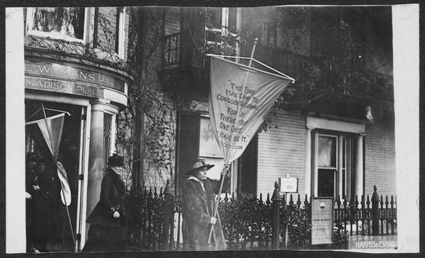 Photograph of Alice Paul emerging from National Woman's Party headquarters holding banner, followed by Dora Lewis (with no banner). Unidentified women stand with a banner in doorway of building. Banner Paul carries reads: "The Time Has Come to Conquer or Submit, For Us There Is But One Choice. [I] Have Made It. President Wilson." Paul is leading picket line from headquarters to the White House.