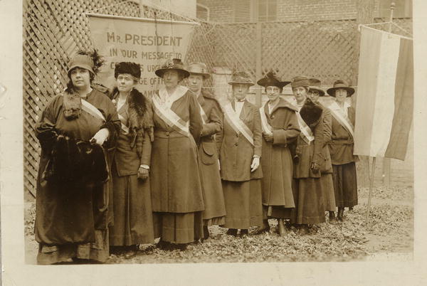 Photograph of nine suffrage pickets standing single file along a tall lattice fence, with suffrage banners. Left to R: Catherine Martinette, Elizabeth Kent, Mary Bartlett Dixon, C. T. Robertson, Cora Week, Amy Jungling, Hattie Kruger, Belle Sheinberg, Julia Emory.
