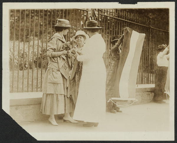 Florence Youmans of Minnesota (left), clutching a suffrage propaganda banner, and Annie Arniel of Delaware (center), being approached in front of the White House gates by an unidentified policewoman, who appears to have seized Arniel's banner, while a third unidentified suffrage picket watches from behind her tri-color purple, white, and gold National Woman's Party flag, and a fourth picket looks away in a different direction.