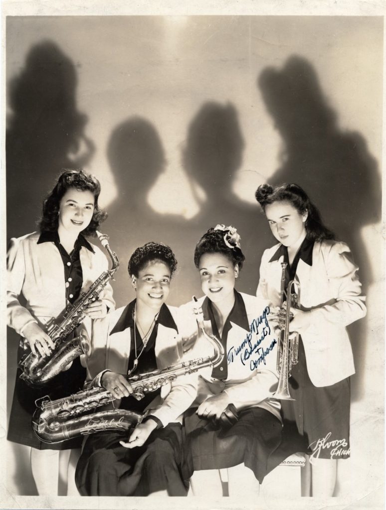 Four beautiful dark skinned women in black skirts and blouses with white blazers hold saxophones and trumpets.