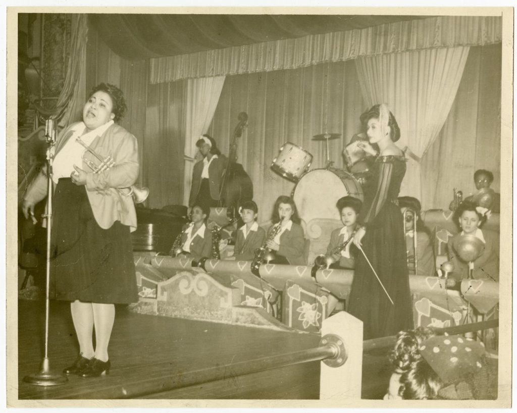 A woman on stage holds a trumpet and sings while a second woman conducts an orchestra of women.