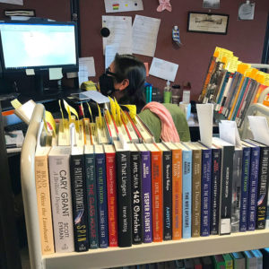 A cataloger sitting at a desk behind a book truck full of books, wearing a mask.
