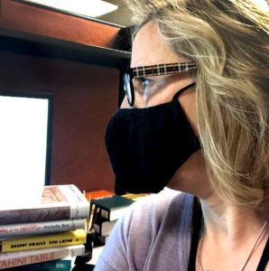 Close up photo of a woman wearing a mask, with a stack of books next to her.