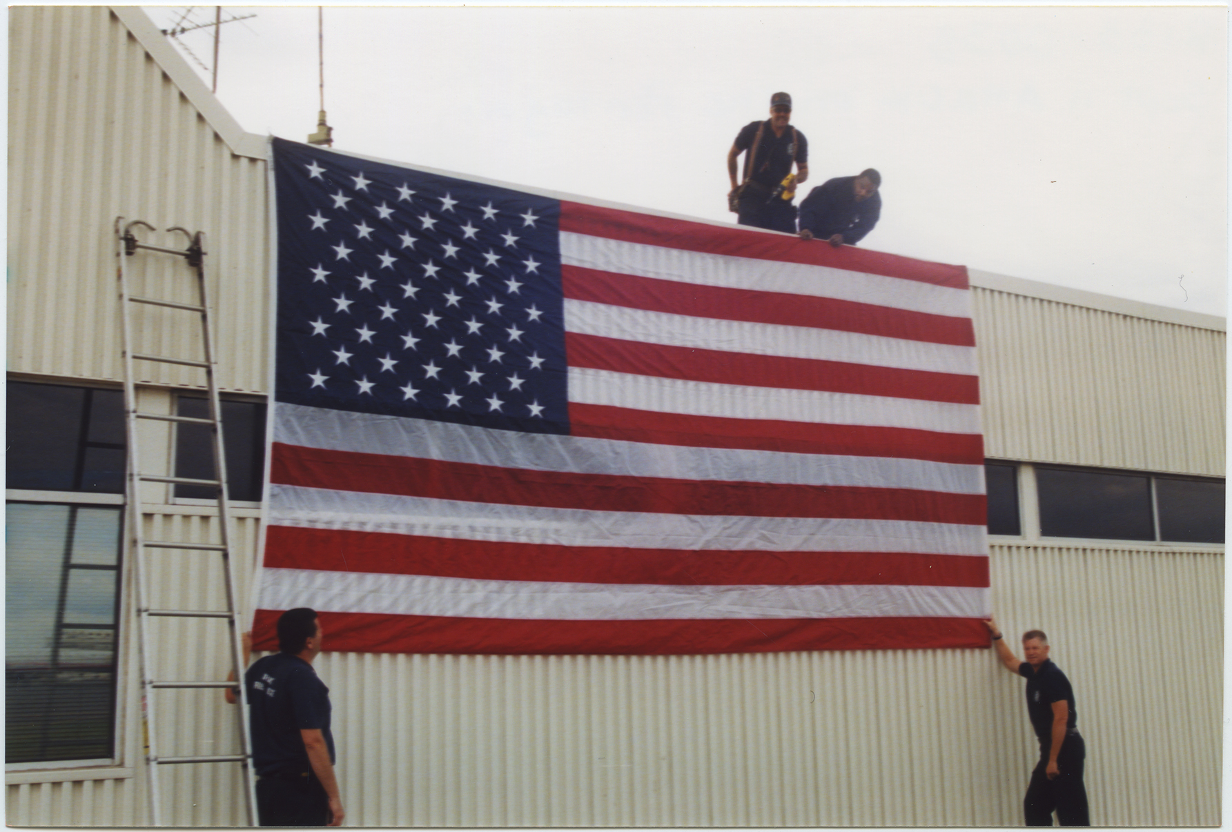 Fire fighters display a large American flag on the front of the National Airport fire station, two days after the 9/11 attack on the Pentagon. 2001, 1 print, col., 4 x 6 in..