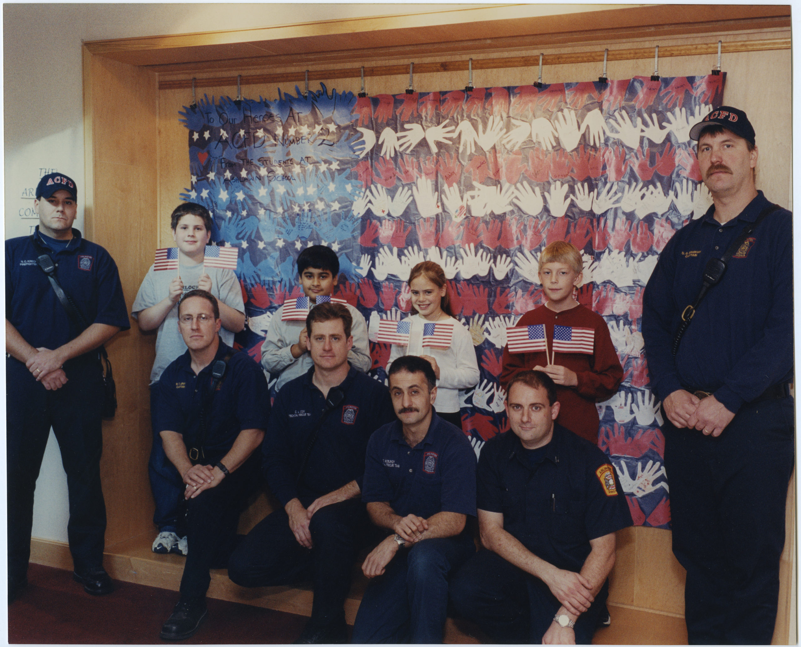 Members of the Arlington County Fire Department posing with children in front of a mural created to thank them for their service.  2001, 1 print, col., 8 x 10 in..