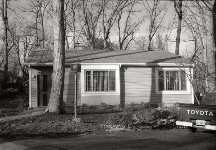 Black and white photo of a one story house with two large square windows and a door on the left side.