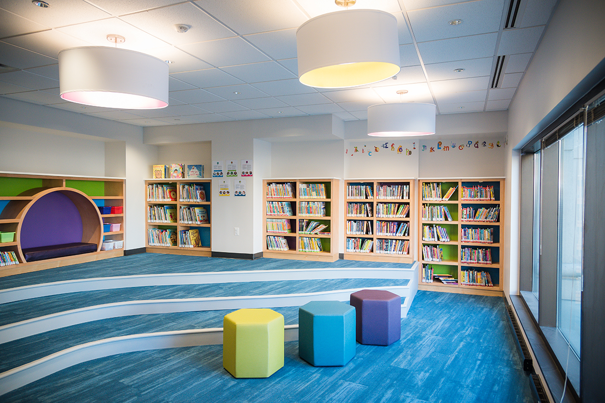 The children's storytime and reading area at Courthouse Library, with three colorful hexagon-shaped seats, a cushioned seating nook, a floor with multiple levels for seating, shelves filled with books and toys, and alphabet decorations on the wall.