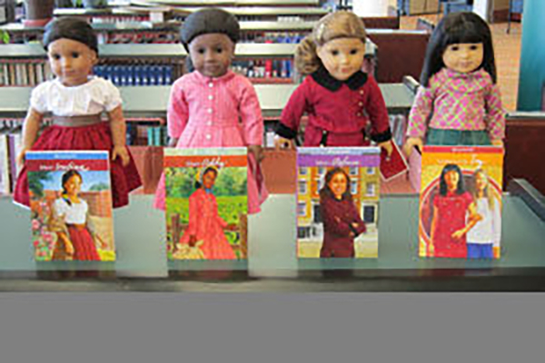 Four American girl dolls stand side-by-side behind their related books.