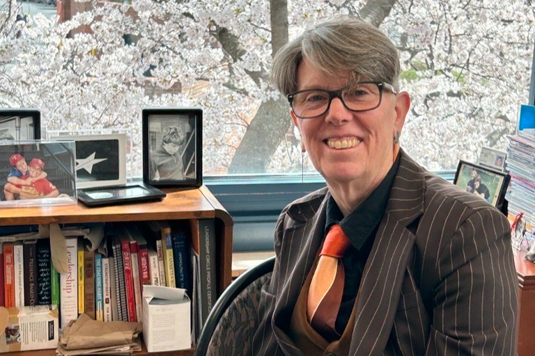 Library Director Diane Kresh smiles at her desk, with a bookshelf and cherry blossoms outside visible in the background.