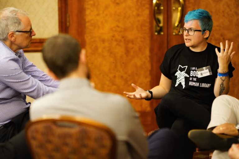 A person with blue hair and a partly visible tattoo wearing a shirt that reads "I am a book of the Human Library" speaks with several people.