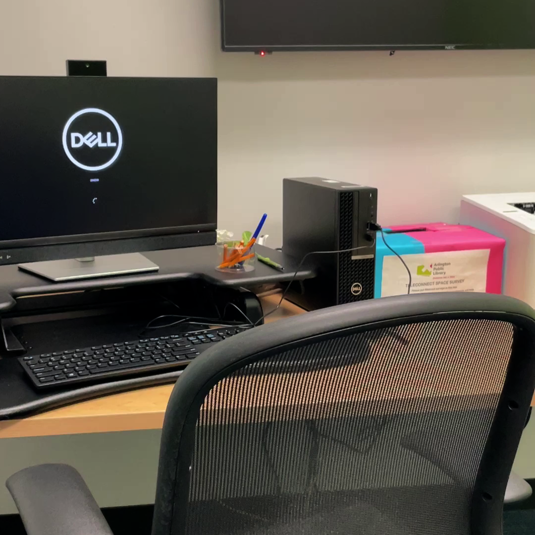 The Teleconnect Space at Columbia Pike featuring a chair, Dell desktop computer with webcam, an adjustable sitting/standing desk, and writing utensils.