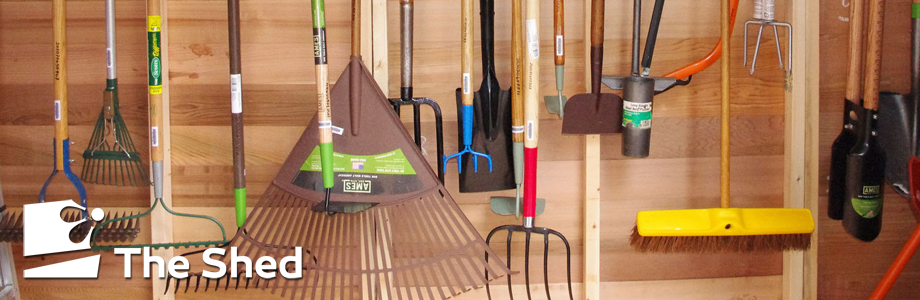 Various garden tools hang against the wall of a shed, including rakes, cultivators, shovels, and more