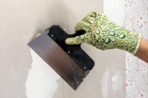 A gloved hand with a putty knife shapes a drywall repair.