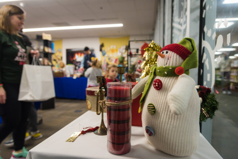 A cozy holiday-themed room with a festive display table in the foreground, featuring a small snowman decoration wearing a red hat and green scarf, alongside a red candle and a 'Peace' ornament. In the background, shoppers browse through various items at a holiday gift nook, with blurred figures adding a warm, bustling atmosphere. A faux fireplace and seasonal decorations add to the festive ambiance.