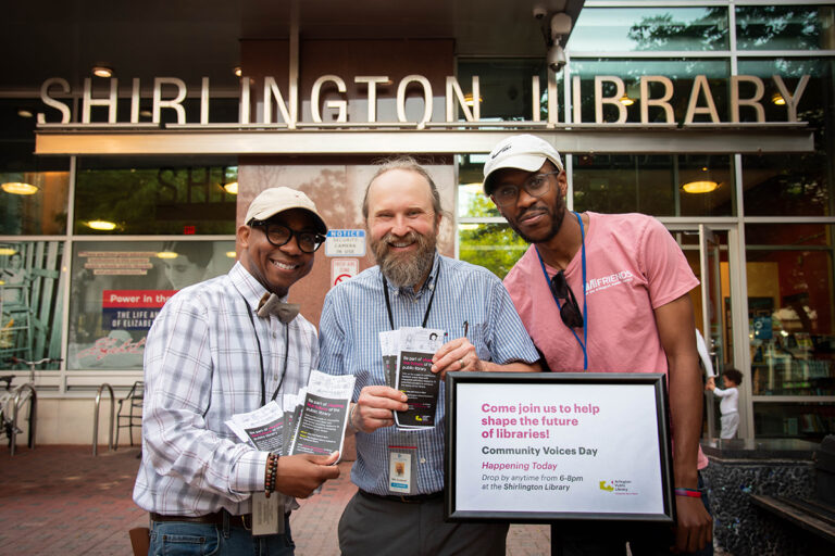 Three Arlington Public Library employees smile outside of Shirlington Library, holding "Be part of shaping the future of Arlington Public Library" pamphlets and standing next to a "Community Voices Day" sign.