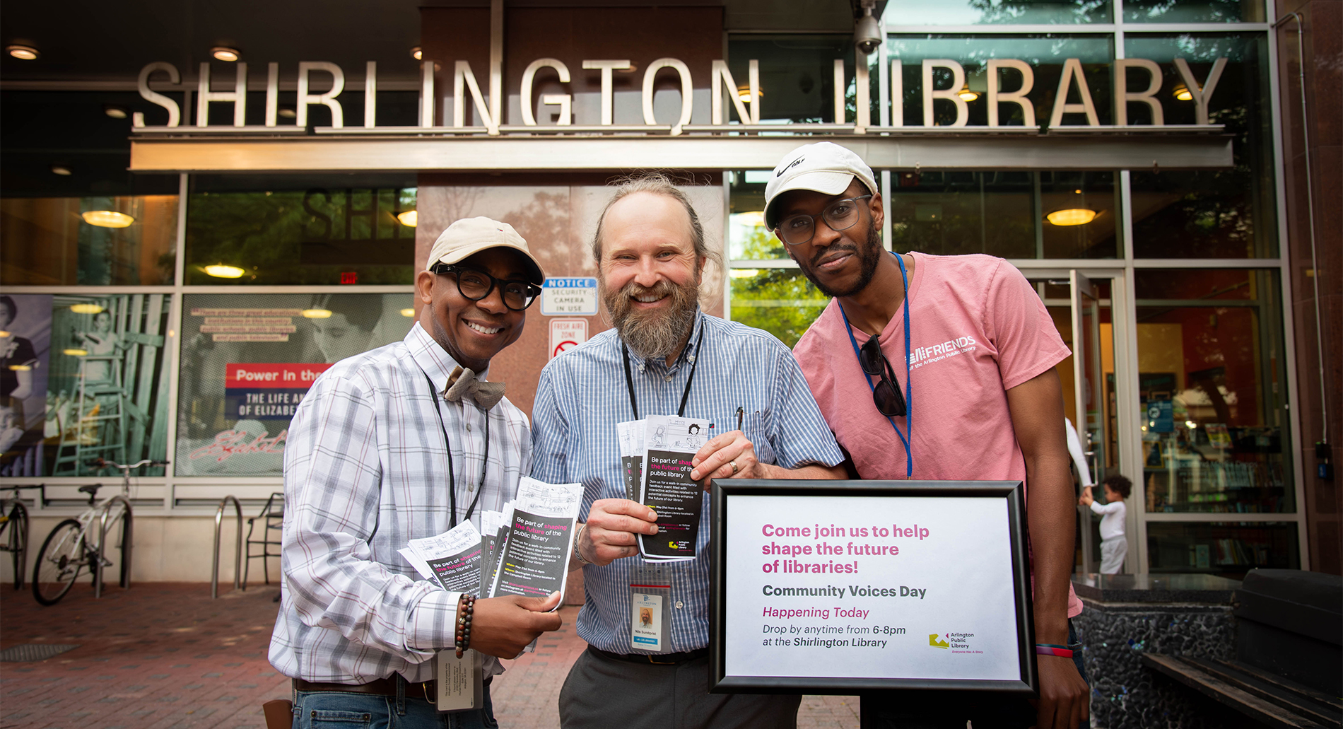 Three Arlington Public Library employees smile outside of Shirlington Library, holding "Be part of shaping the future of Arlington Public Library" pamphlets and standing next to a "Community Voices Day" sign.