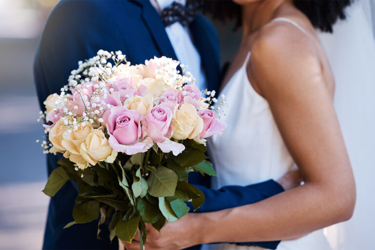 A couple standing close together outdoors, with one holding a bouquet of pink and cream roses accented with baby's breath. One person is dressed in a navy suit and bow tie, and the other is wearing a white gown with thin straps.