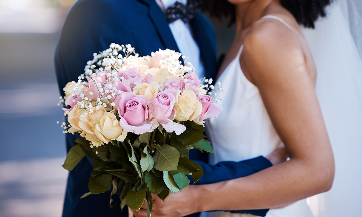 A couple standing close together outdoors, with one holding a bouquet of pink and cream roses accented with baby's breath. One person is dressed in a navy suit and bow tie, and the other is wearing a white gown with thin straps.