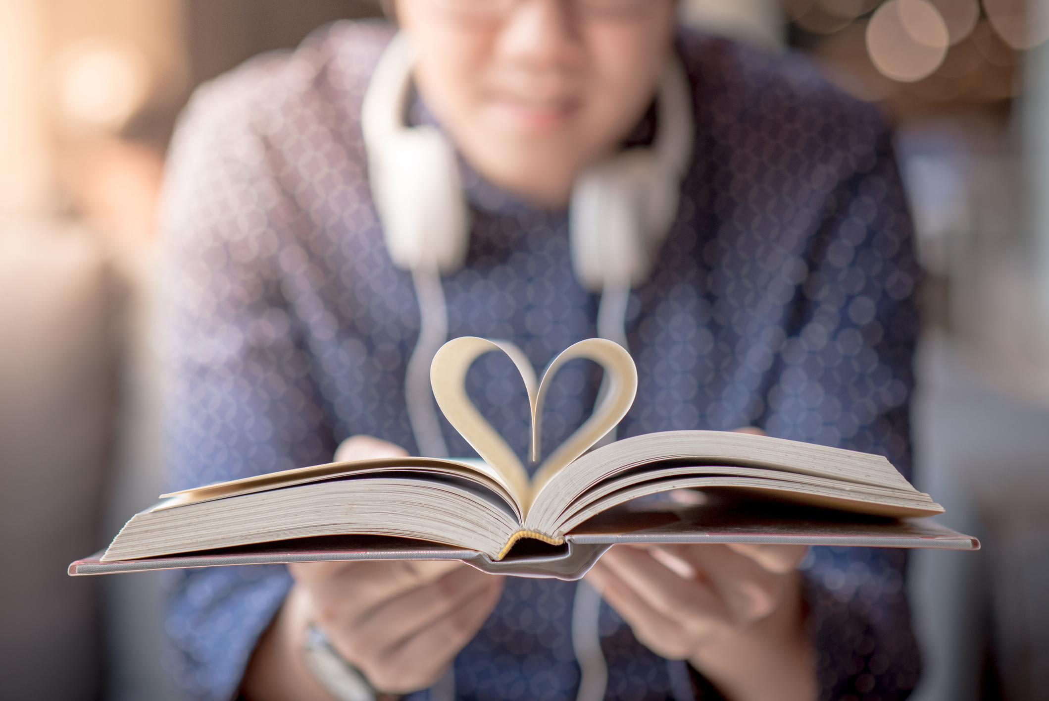 A close-up of an open book with its pages folded to form a heart shape in the center, held by a person wearing a patterned shirt and headphones around their neck. The background is softly blurred, creating a warm, cozy atmosphere.