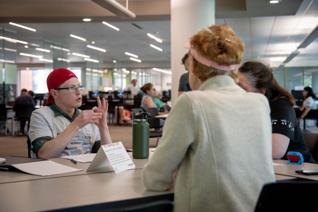 Deaf Plus Advocate Jeremy Smith, wearing a red cap and glasses, signs expressively in conversation with a participant during Arlington Public Library's Human Library event. A sign on the table identifies his topic, 'My DeafPlus Life,' as part of the program. The library's open and modern space is visible in the background, with other participants seated at tables.