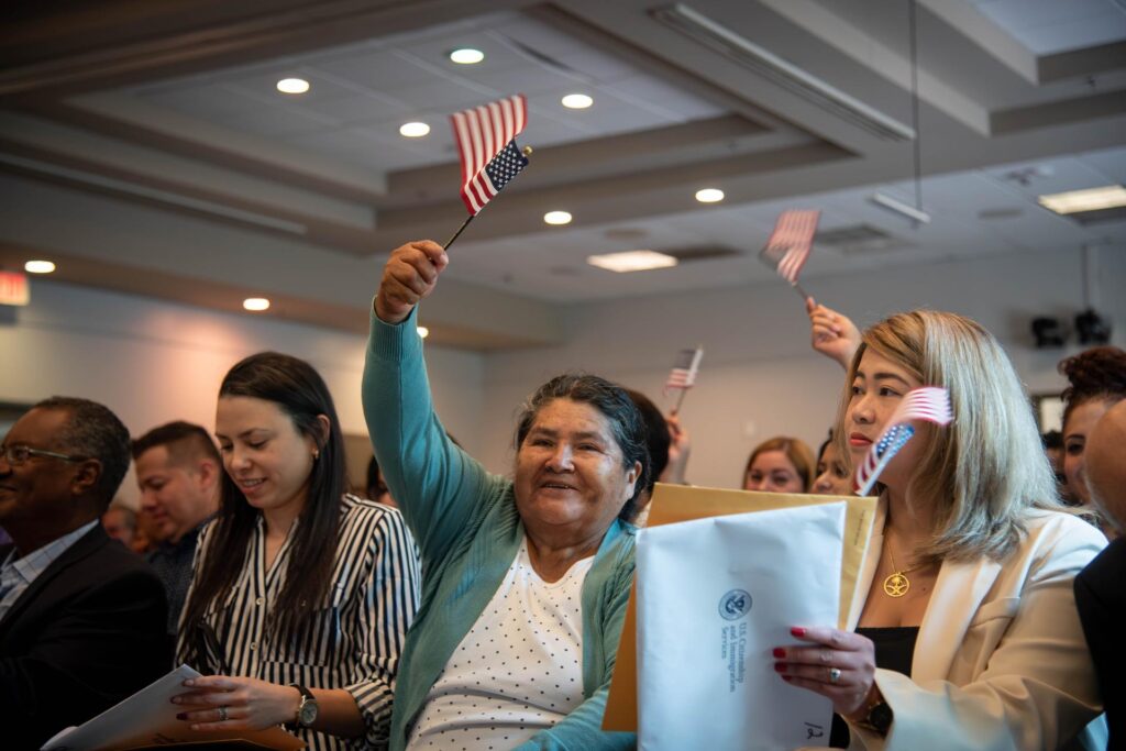 A joyful moment during a Naturalization Ceremony at Central Library, with a woman in a teal cardigan proudly waving a small American flag. She is surrounded by other participants, some holding flags and official documents from U.S. Citizenship and Immigration Services, all celebrating their new citizenship.