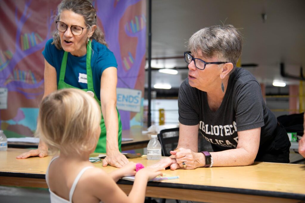 Library Director Diane Kresh leans over a table in conversation with a young girl during a book sale in the Central Library parking garage. Another library volunteer wearing a green apron is engaged nearby. The table is surrounded by books and event signage, with a colorful backdrop featuring illustrations of books and trees.