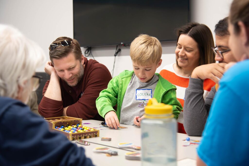 A group of participants enjoys a board game at Arlington Public Library's GameFest event. A young boy wearing a green hoodie focuses intently on his game piece while an adult next to him, smiling and wearing sunglasses on his head, watches attentively. Other players, including a woman in a striped sweater and additional participants blurred in the foreground, are engaged in the activity. A colorful game setup with dice and cards is spread across the table, creating a fun and interactive atmosphere in the library space. A mounted TV screen is visible in the background.