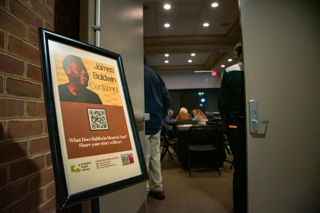 A photograph of a James Baldwin community read event at Central Library. In the foreground, a framed poster promotes the "James Baldwin Centennial," featuring an image of Baldwin smiling against a warm-toned background. Below, text invites attendees to share their stories, with a QR code and the Arlington Public Library logo displayed. The poster is positioned next to an open door leading into a well-lit room where participants are seated at tables, engaged in discussion. The atmosphere is warm and inviting, with visible details of the brick wall and modern interior lighting.