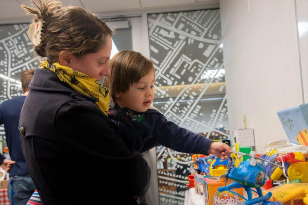 A mother and young child explore a table filled with colorful toys at Arlington Public Library's Holiday Gift Nook. The mother, wearing a yellow scarf with a pattern, holds the child as they reach out toward a blue toy helicopter. The background features a frosted glass wall with a map-like design, adding a festive yet modern ambiance to the event. The table is stocked with various toys and games, creating a cheerful and family-friendly atmosphere.