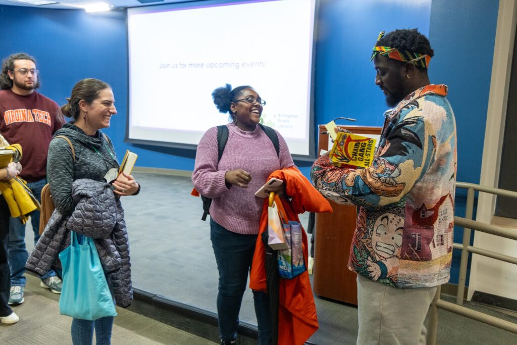 Author Nana Kwame Adjei-Brenyah signs books and chats with attendees after his Arlington Reads event. He is wearing a colorful jacket and headband, holding a stack of books, including one titled "Chain-Gang All-Stars." A group of smiling attendees surrounds him, holding books and coats. The background shows a blue wall and a large projector screen displaying the Arlington Public Library logo and text encouraging participation in future events. The atmosphere is lively and engaging.