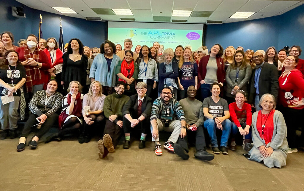 A large group photo of Arlington Public Library staff gathered at a holiday party. The group is diverse, with attendees dressed in festive attire, including holiday sweaters, bright scarves, and red accents. Some individuals wear name tags, and a few in the front row are seated on the floor, smiling cheerfully. In the background, a presentation screen displays text about the upcoming APL Trivia Tournament, set for Spring 2025. The atmosphere is warm and joyful, showcasing camaraderie and celebration among the staff.