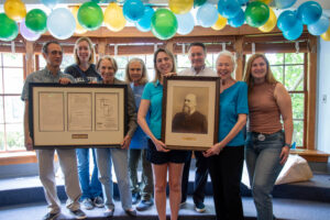 A group of eight people stands together on a small stage in front of large windows, smiling at the camera. They hold two framed items—one is a historical document with maps and text, and the other is a framed portrait of a bearded man. Colorful blue, green, and yellow balloons decorate the space above them, celebrating Glencarlyn Library's 101st birthday.