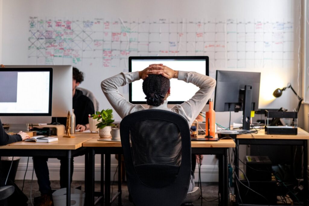 A modern office setting with multiple employees working at their desks. In the foreground, a person with dark, curly hair tied back sits in a black mesh office chair, facing a computer screen with their hands resting on the back of their head.