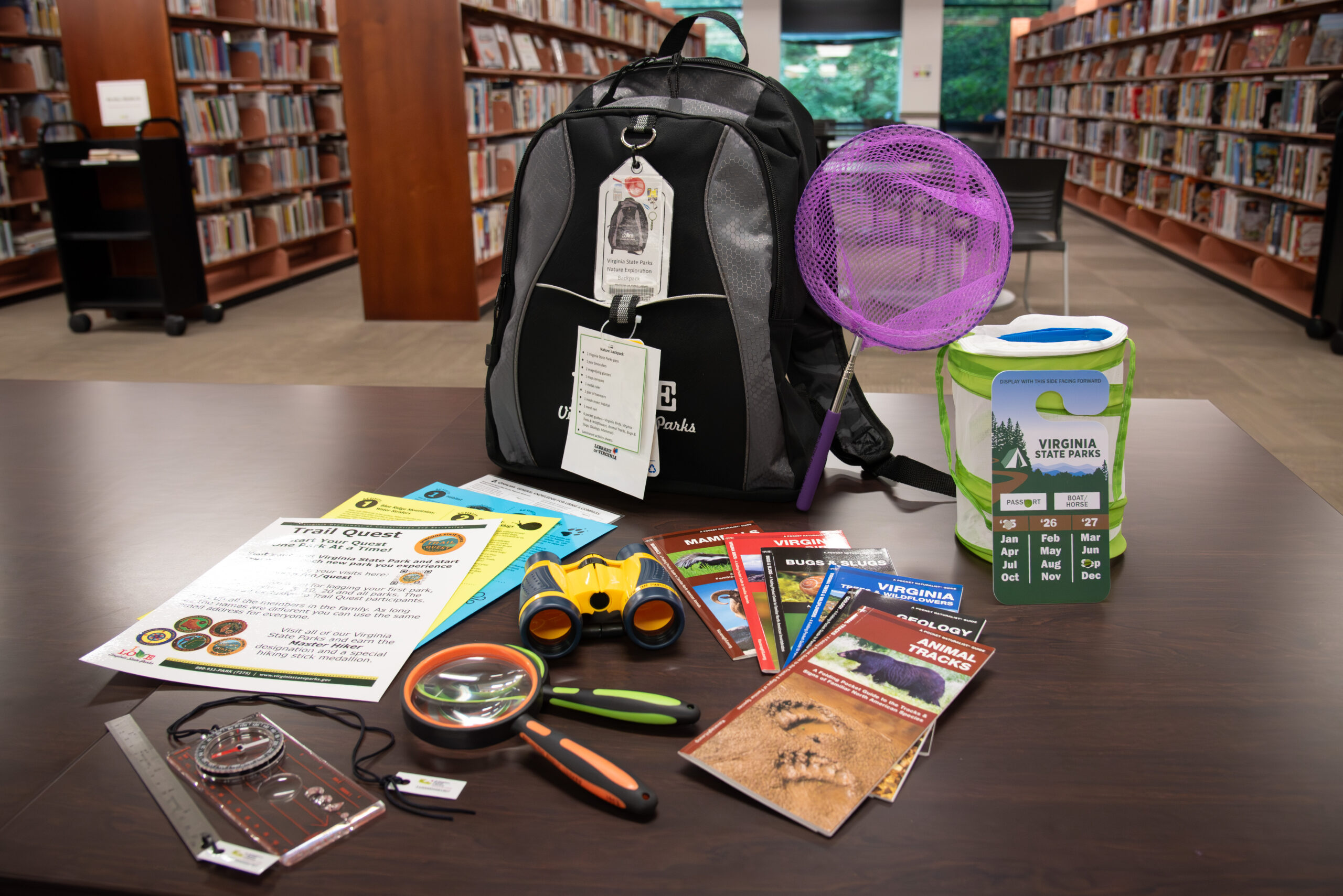 A Nature Backpack from Arlington Public Library is displayed on a table in the library, with bookshelves in the background. The black and gray backpack is surrounded by educational tools and activity guides, including a purple bug net, a portable mesh butterfly habitat, a green Virginia State Parks pass, binoculars, a compass, magnifying glass, tweezers, and various wildlife and trail brochures. A laminated checklist and “Trail Quest” activity sheet are also visible, promoting exploration of Virginia’s state parks.