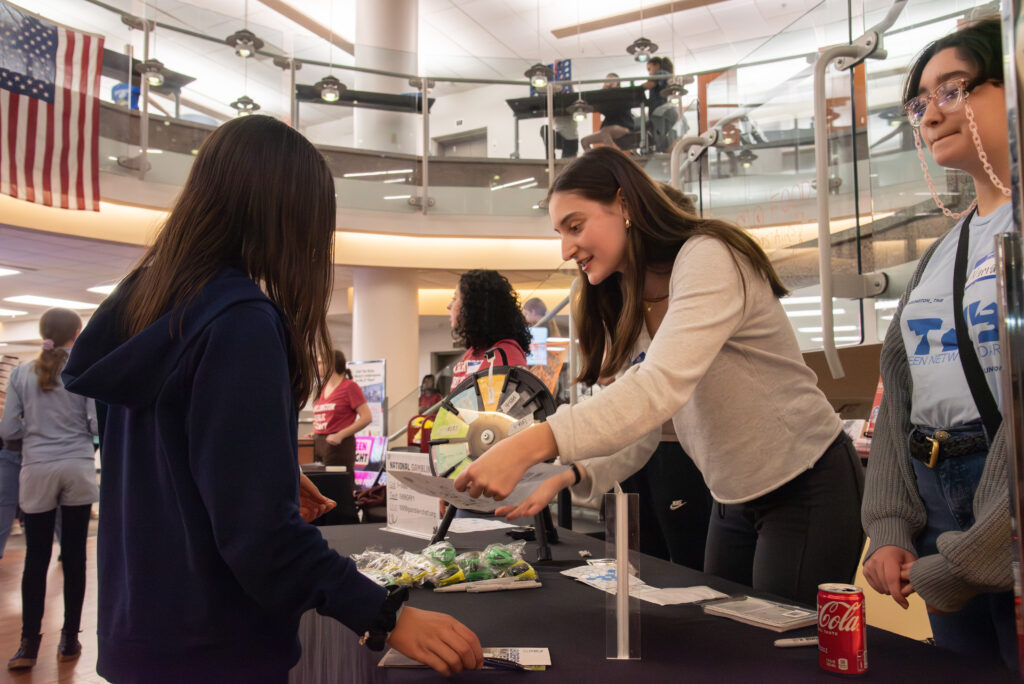 A teen volunteer welcomes a younger teen to a table with a spinning wheel game at Central Library.