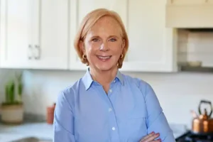 Anne Byrn smiles with her arms crossed, standing in a bright kitchen while wearing a bright blue button-down.