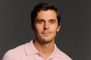 Antoni Porowski smiles, wearing a pink shirt in front of a dark gray background.