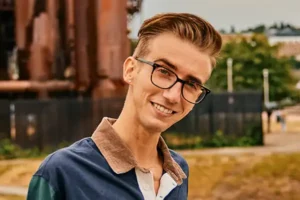 Benjamin Dylan Hollis smiles outside in front of a metal structure, wearing a collared jacket and glasses.