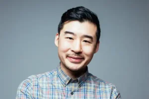 Francis Lam smiles wearing a plaid shirt against a gray background.