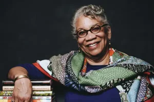 Jessica B. Harris smiles while leaning on a stack of books, wearing a multipatterned shawl and glasses.