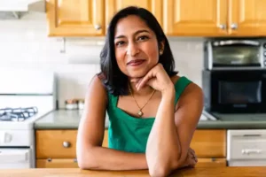 Author Priya Krishna smiles while leaning on a kitchen counter, with a stove and microwave visible in the background.