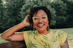 Dr. Ashanté M. Reese sitting on a wooden bench outdoors, wearing a yellow patterned top with greenery in the background.