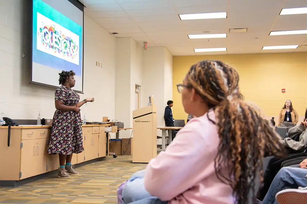 Author Roseanne A. Brown presents to a class of in front of a screen with text that reads: Questions?