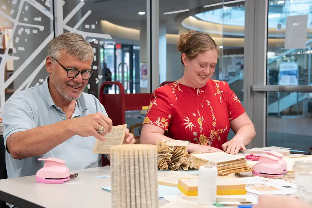 Two people sit at a table inside Arlington Public Library, folding pages and crafting paper decorations for the wedding event.