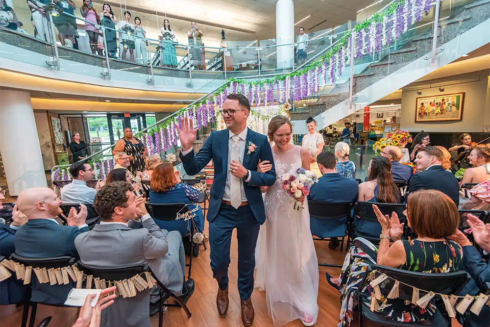 A newly married couple walks arm in arm down the aisle at Central Library, greeted by applause from seated guests and surrounded by festive decorations.